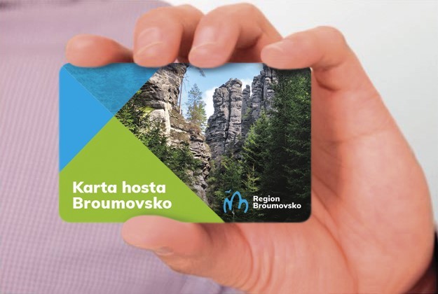 Ruka drží turistickou kartu s fotografií skal a logem Broumovska, na modro-zeleném pozadí.